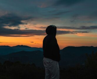 Persoon in donkere hoodie kijkt uit over bergen bij een kleurrijke zonsondergang